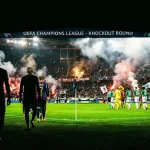 Football players emerging from a tunnel into a packed stadium under bright lights for a knockout round match